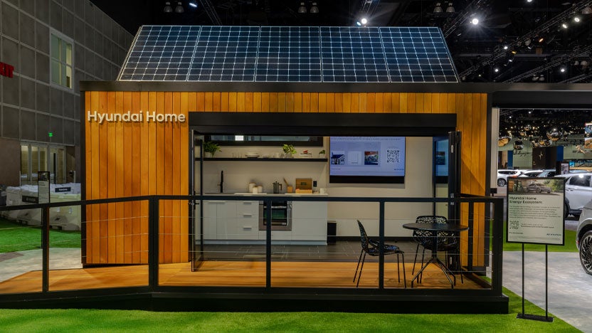 A single-story house with solar panels on its dark gray roof. The house has light-colored walls and is surrounded by lush green bushes and trees. A paved driveway leads to the house.